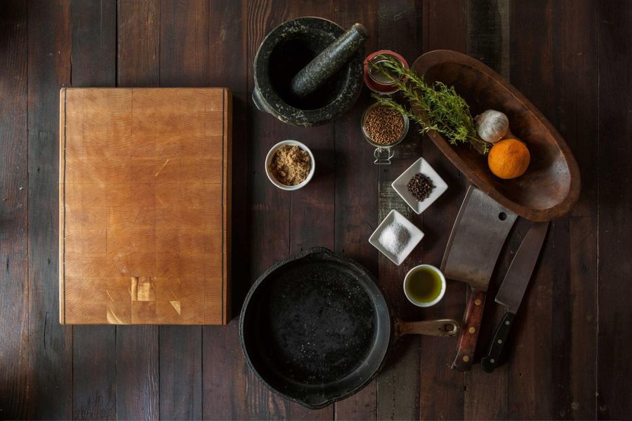 End-grain maple chopping board on dark plank floor beside cast-iron pan and stone mortar