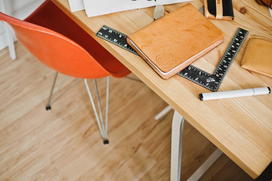 Light wooden table edge with a leather notebook, steel rules and an orange chair beyond