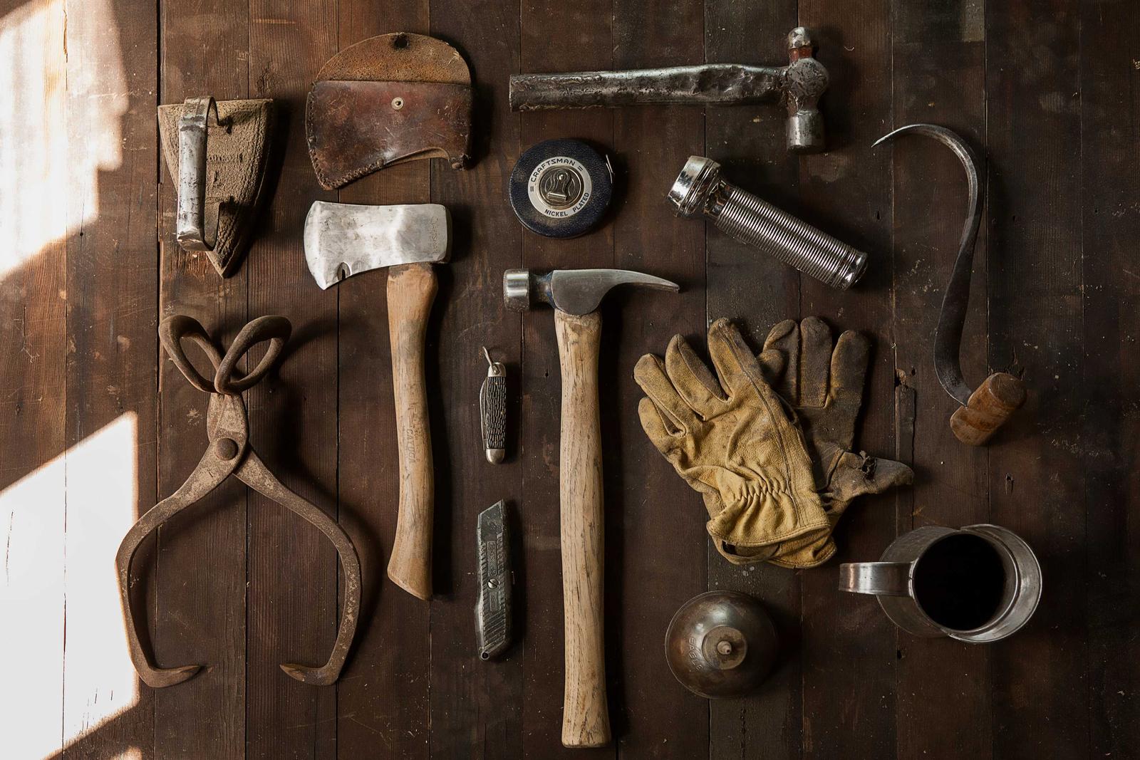 Old hand tools laid out on dark floorboards: axe, hammers, ice tongs, leather gloves and a tape measure, lit from the left